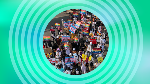 A crowd marches in Myanmar with rainbow flag and trans flag signs reading #LGBTQ4Democracy